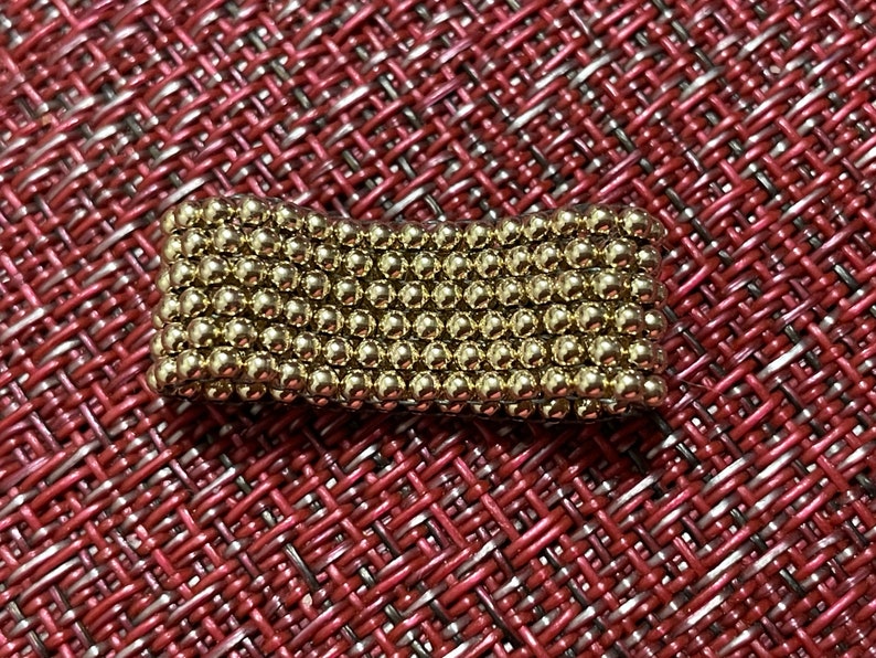 May include: A close-up of a gold-coloured magnetic ball chain. The chain is made up of small, round balls that are connected together. The chain is lying on a red and white patterned surface.
