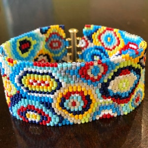 Puede incluir: Un brazalete de cuentas de colores con un patrón geométrico de círculos y espirales en tonos de azul, rojo, amarillo, blanco y plateado.