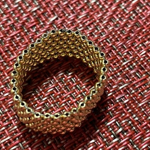 May include: A gold-toned ring made of small, round beads. The ring is on a red and white textured surface.