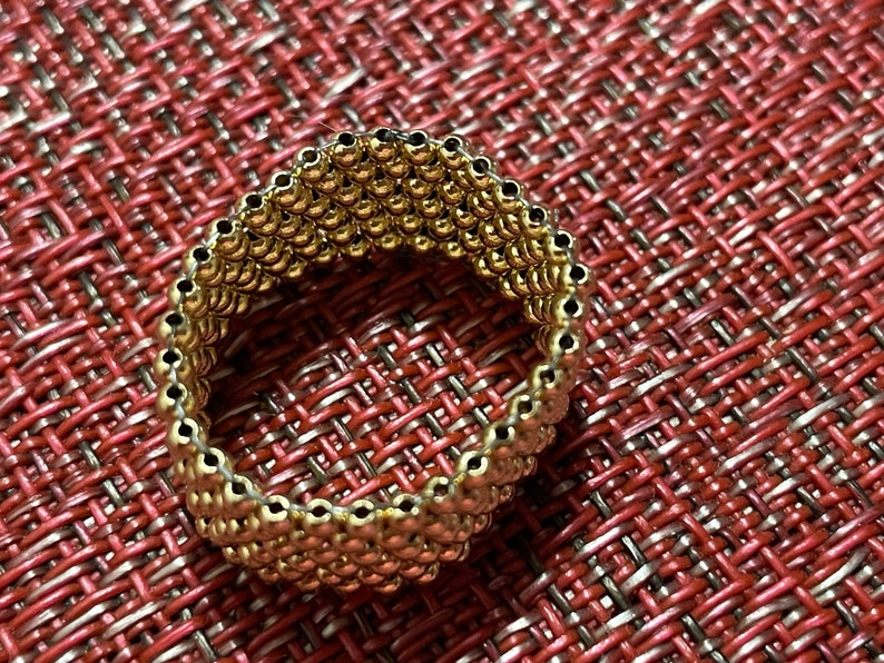 May include: A gold-toned ring made of small, round beads. The ring is on a red and white textured surface.
