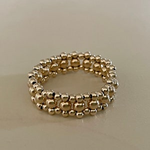 Puede incluir: Un anillo dorado compuesto por múltiples filas de pequeñas cuentas esféricas. El anillo es circular y se asienta sobre una superficie de tonos neutros, mostrando su acabado metálico reflectante. El diseño es simple pero elegante.