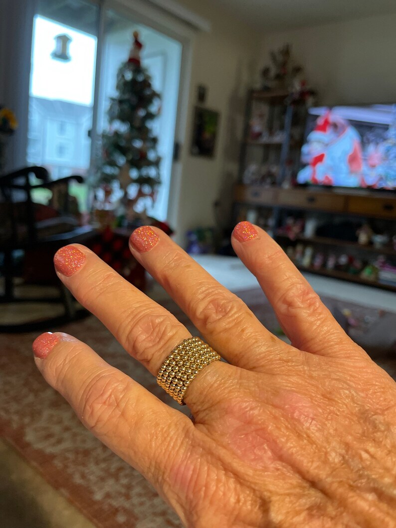May include: A gold beaded ring with multiple rows of beads. The ring is worn on a hand with pink glitter nail polish.