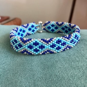 May include: A blue, grey, and light blue beaded bracelet with a geometric pattern.