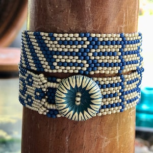 May include: A beaded bracelet featuring a blue and silver geometric design. The bracelet is fastened with a large, round button with a blue and cream pattern. The bracelet is wrapped around a wooden post.