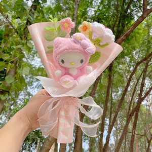 May include: A pink Hello Kitty plush toy is arranged in a bouquet of pink and white flowers. The bouquet is wrapped in pink and white checked paper and tied with a pink ribbon.