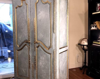 May include: A tall, light blue armoire with a distressed finish and gold-painted trim. The two-door cabinet features ornate gold molding, carved shell details at the top and bottom, and vertical gold hardware handles on the center doors.