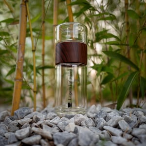 May include: A clear glass bottle with a wooden lid and a black label that says "Lagom Infusions". The bottle is sitting on a bed of small gray rocks.