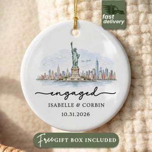 May include: A round white ceramic ornament featuring a watercolor illustration of the Statue of Liberty and the New York City skyline. The text reads engaged, Isabelle & Corbin, and 10.31.2026. It hangs from a gold string against a textured background.