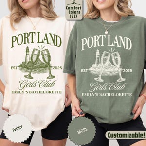 May include: Two short-sleeve t-shirts in ivory and moss green featuring a graphic print with the text PORT LAND, EST 2025, Girls Club, and EMILY'S BACHELORETTE. The design includes a lighthouse and two champagne flutes clinking together.