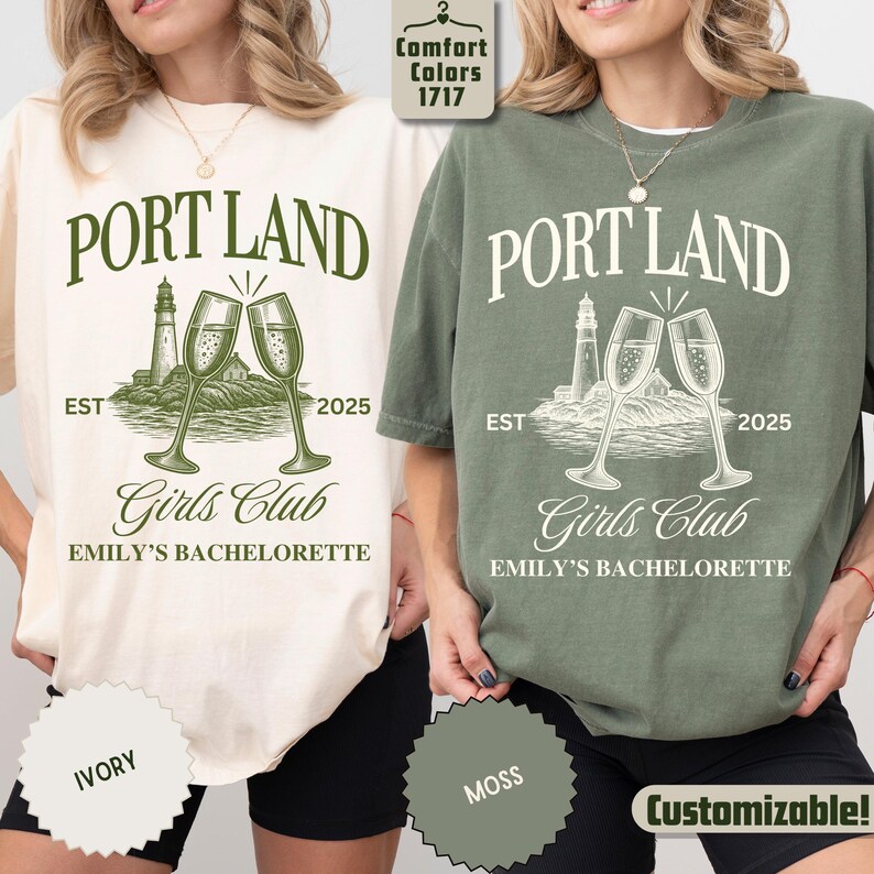 May include: Two short-sleeve t-shirts in ivory and moss green featuring a graphic print with the text PORT LAND, EST 2025, Girls Club, and EMILY'S BACHELORETTE. The design includes a lighthouse and two champagne flutes clinking together.