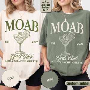 May include: Two t-shirts, one ivory and one moss green, feature the word "MOAB" above a cocktail illustration. Below the graphic are the words "Girls Club" and "Emily's Bachelorette". The year "2025" is also printed on the shirts.