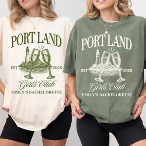 May include: Two oversized t-shirts, one cream and one olive green, featuring a graphic print with the text PORT LAND, EST 2025, Girls Club, and EMILY'S BACHELORETTE. The design includes a lighthouse on a rocky shore and two clinking champagne flutes.