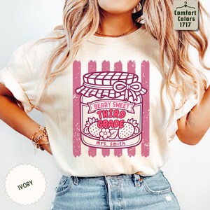 May include: An ivory t-shirt featuring a graphic of a jam jar with a checkered lid and bow. The jar contains strawberries and the text "BERRY SWEET THIRD GRADE" and "mrs. smith" in pink. The design is set against vertical pink stripes.