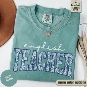 May include: A light green Comfort Colors t-shirt with the words "english TEACHER" in white and blue floral print. The shirt has a crew neck and short sleeves. A silver necklace with a pendant is draped across the shirt.
