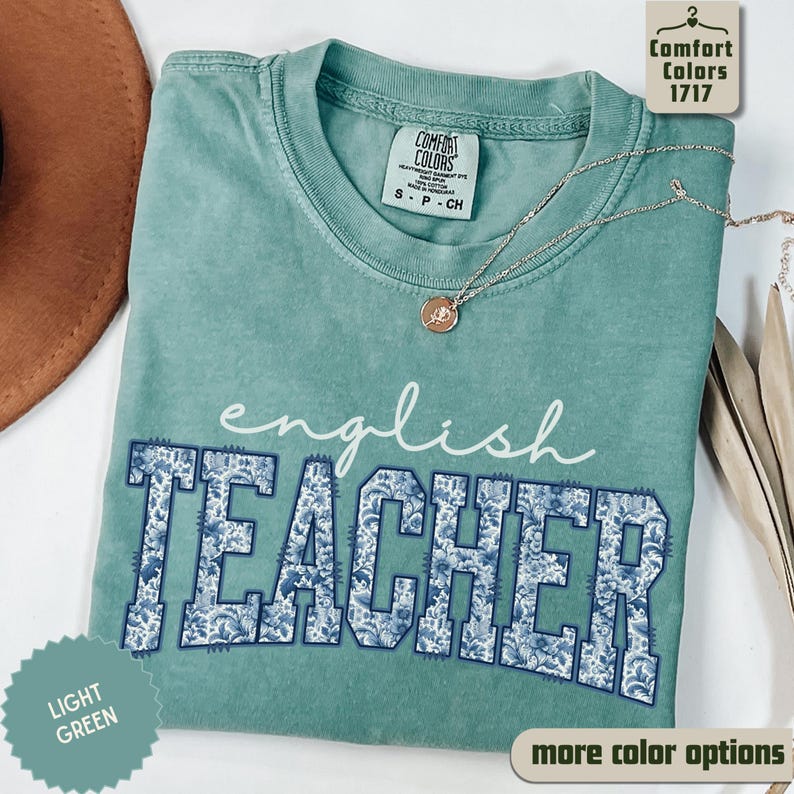 May include: A light green Comfort Colors t-shirt with the words "english TEACHER" in white and blue floral print. The shirt has a crew neck and short sleeves. A silver necklace with a pendant is draped across the shirt.