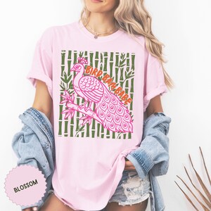 Może przedstawiać: Jasnor&oacute;żowy t-shirt z grafiką przedstawiającą r&oacute;żowego pawia siedzącego na gałęzi bambusa. Tło składa się z zielonych łodyg bambusa. Napis BIRD BAM BABE jest wydrukowany na pomarańczowo. Mała okrągła metka zawiera napis BLOSSOM.