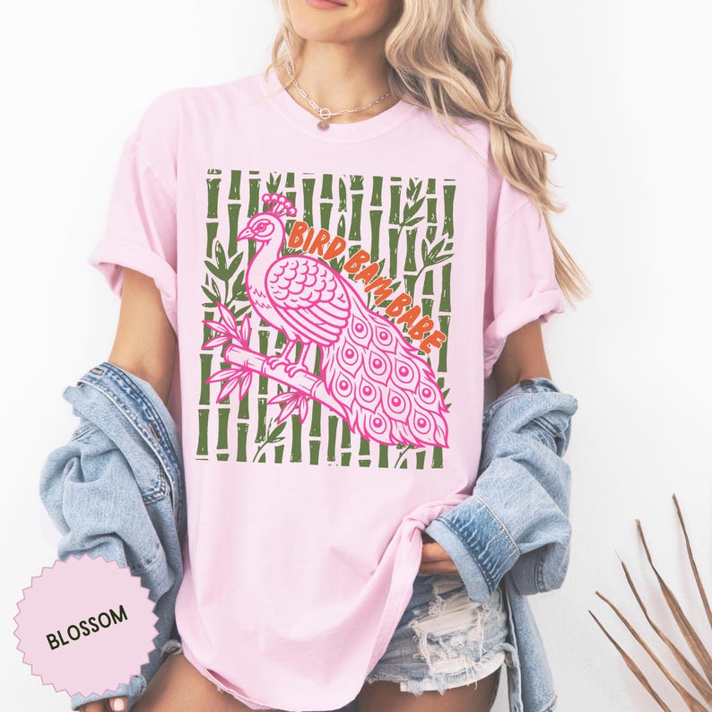 Może przedstawiać: Jasnor&oacute;żowy t-shirt z grafiką przedstawiającą r&oacute;żowego pawia siedzącego na gałęzi bambusa. Tło składa się z zielonych łodyg bambusa. Napis BIRD BAM BABE jest wydrukowany na pomarańczowo. Mała okrągła metka zawiera napis BLOSSOM.