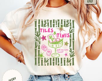 Op de afbeelding: Een ivoorkleurig t-shirt met een grafische print met een bamboerand, de tekst "TILES & TINIS" in het roze, een martiniglas met garnering, mahjongtegels en de tekst "just one more round!" in het roze, geaccentueerd met groene en roze sterren.