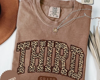 May include: A brown Comfort Colors t-shirt with the word "THIRD" in a leopard print design. The word "GRADE" is below the word "THIRD". A silver necklace with a pendant is on the shirt.