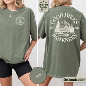 May include: Olive green t-shirt with the text "SCENIC & SIXTY YELLOWSTONE" on the front and a graphic on the back that says "GOOD HIKES BAD KNEES" with a cabin and mountain design. The shirt is a comfort color and is customizable.