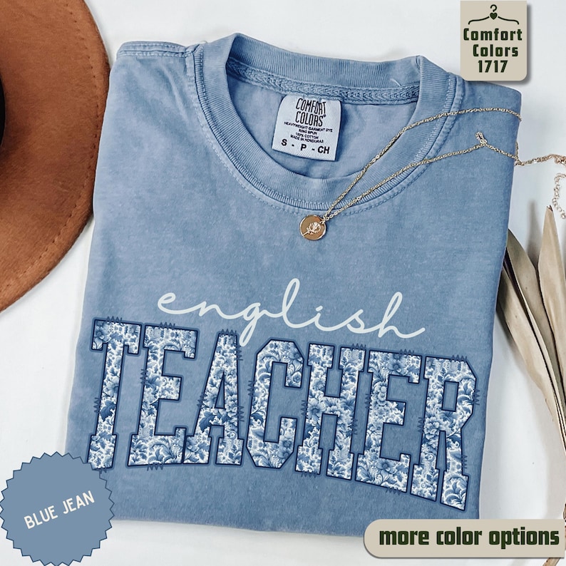 May include: A blue jean Comfort Colors t-shirt with the words "english TEACHER" in a floral design. The shirt has a crew neck and short sleeves. A gold necklace with a pendant is draped on the shirt.