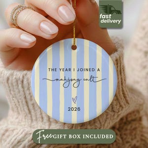 May include: A round ceramic ornament featuring vertical light blue and cream stripes. The text reads THE YEAR I JOINED A mahjong cult, followed by a small purple heart icon and the year 2026. A gold string is attached to the top for hanging.