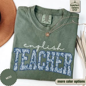 May include: A moss green t-shirt with the words "english TEACHER" in white and blue floral print. The shirt has a crew neck and short sleeves. A gold necklace with a pendant is draped on the shirt. The shirt is labeled "Comfort Colors 1717".