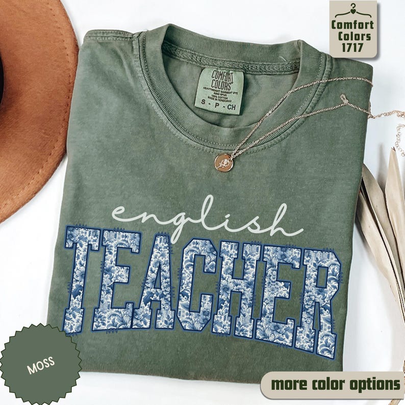 May include: A moss green t-shirt with the words "english TEACHER" in white and blue floral print. The shirt has a crew neck and short sleeves. A gold necklace with a pendant is draped on the shirt. The shirt is labeled "Comfort Colors 1717".