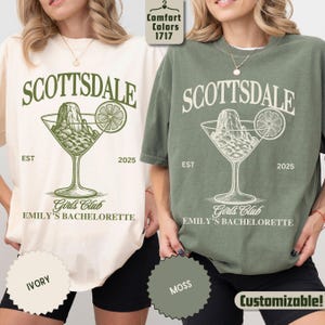 May include: Two t-shirts, one ivory and one moss green, featuring the word "SCOTTSDALE" above a cocktail glass design with a mountain and lime slice. The text "EST 2025" and "Girls Club EMILY'S BACHELORETTE" are also printed on the shirts.