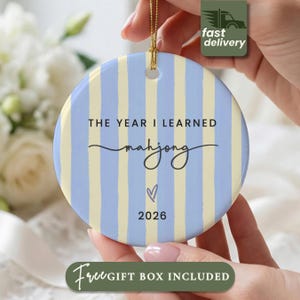 May include: A round ceramic ornament featuring vertical light blue and cream stripes. The text reads THE YEAR I LEARNED mahjong with a small heart icon and the year 2026 printed in black. A gold string is attached to the top for hanging.