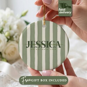 May include: A round ceramic ornament featuring vertical sage green and white stripes. The center displays the name JESSICA in bold dark green serif lettering, with the text occupational therapist and ESTABLISHED 2026 printed below it.