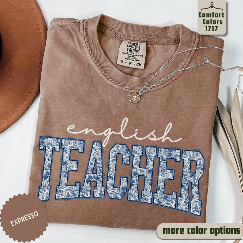 May include: A brown t-shirt with the words "english TEACHER" printed on the front. The word "english" is in a cursive font, and "TEACHER" is in a block letter style with a blue floral pattern. A silver necklace is draped across the shirt.
