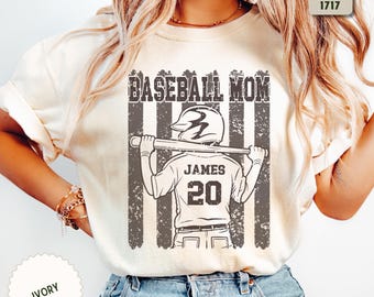 Może przedstawiać: Koszulka w kolorze kości słoniowej z ciemnoszarą grafiką przedstawiającą baseballistę w kasku z kijem. Napis głosi BASEBALL MOM, JAMES oraz numer 20. Etykieta wskazuje Comfort Colors 1717 oraz kolor kości słoniowej.