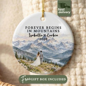 May include: A round white ceramic ornament featuring a printed mountain landscape with a lake and a couple standing in the foreground. The text reads FOREVER BEGINS IN MOUNTAINS, Isabelle and Corbin, and 2026. It includes a gold hanging string.