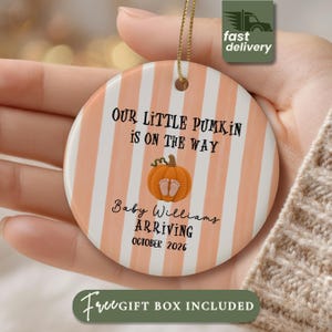 May include: A round ceramic ornament featuring vertical peach and white stripes. The text reads: Our little pumkin is on the way, Baby Williams arriving, October 2026. A graphic of an orange crocheted pumpkin with baby feet is in the center.
