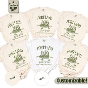 May include: Five short-sleeved crew neck t-shirts in ivory and white. Each shirt features a dark green graphic with the text PORT LAND, EST 2025, Girls Club, and EMILY'S BACHELORETTE, alongside an illustration of a lighthouse and two champagne flutes.