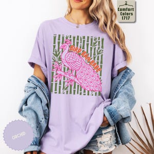 Może przedstawiać: Jasnofioletowy t-shirt z kr&oacute;tkim rękawem z grafiką r&oacute;żowego pawia siedzącego na gałęzi bambusa. Tło grafiki składa się z zielonych łodyg bambusa. Napis BIRD BATH BABE jest wydrukowany pomarańczowymi literami nad pawiem.