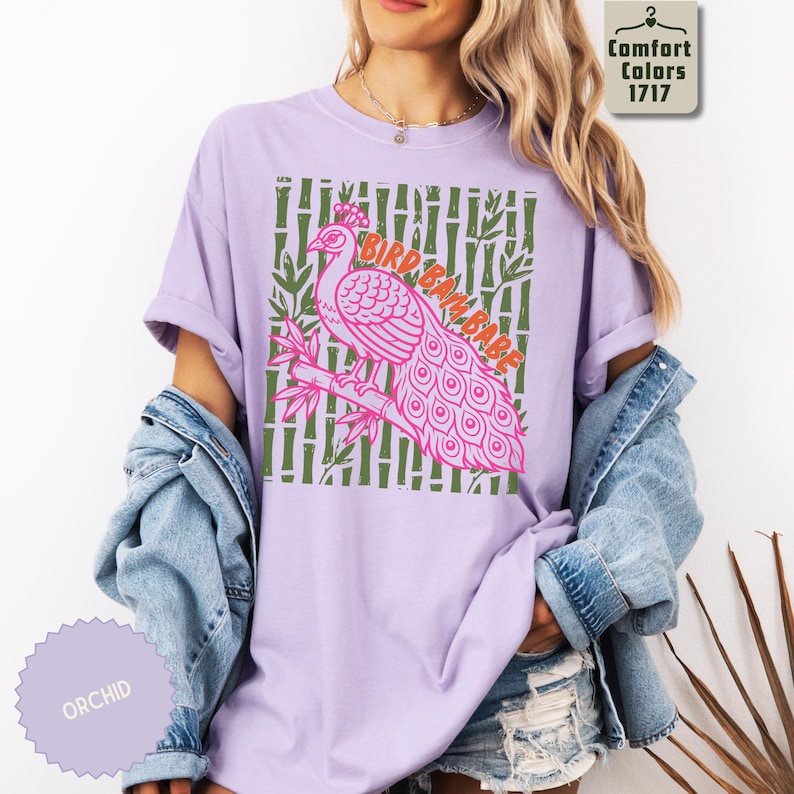 Może przedstawiać: Jasnofioletowy t-shirt z kr&oacute;tkim rękawem z grafiką r&oacute;żowego pawia siedzącego na gałęzi bambusa. Tło grafiki składa się z zielonych łodyg bambusa. Napis BIRD BATH BABE jest wydrukowany pomarańczowymi literami nad pawiem.