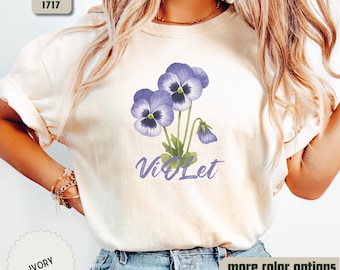 May include: A white t-shirt with a purple flower design and the word "Violet" printed on the front. The shirt is being modeled by a person wearing blue jeans.