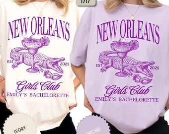 May include: Two t-shirts, one ivory and one orchid, featuring a purple graphic. The graphic includes the text "NEW ORLEANS", an alligator, a cocktail, and the words "Girls Club" and "EMILY'S BACHELORETTE".