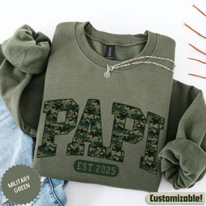 May include: Military green sweatshirt with the word "PAPI" in camouflage print. The text "EST 2025" is below the word "PAPI". A silver necklace with a circular pendant is on the sweatshirt. The sweatshirt is folded on top of blue jeans.