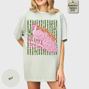 Może przedstawiać: Jasnozielony t-shirt z kr&oacute;tkim rękawem z grafiką r&oacute;żowego pawia siedzącego na gałęzi bambusa. Wz&oacute;r zawiera napis "BIRD BAM BABE" pomarańczowymi literami na tle z motywem ciemnozielonych łodyg bambusa.