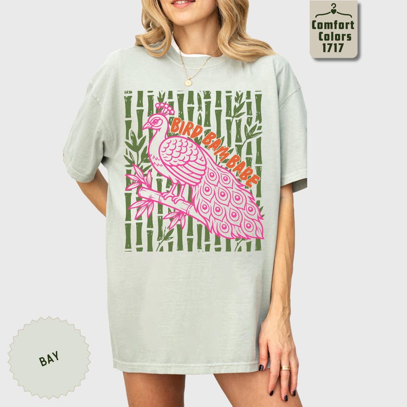 Może przedstawiać: Jasnozielony t-shirt z kr&oacute;tkim rękawem z grafiką r&oacute;żowego pawia siedzącego na gałęzi bambusa. Wz&oacute;r zawiera napis "BIRD BAM BABE" pomarańczowymi literami na tle z motywem ciemnozielonych łodyg bambusa.