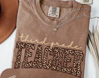 May include: Espresso-colored Comfort Colors t-shirt with the words "third grade TEACHER" printed on the front. The word "TEACHER" is in a leopard print design. A silver necklace with a pendant is draped across the shirt. The shirt is folded.