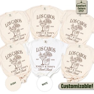 May include: A collection of ivory and white t-shirts with the text "LOS CABOS" and "EMILY & TOM'S Bach Bash" printed in brown. Each shirt features a graphic of a mountain, a margarita, and a cocktail. The text "2025" is also included.