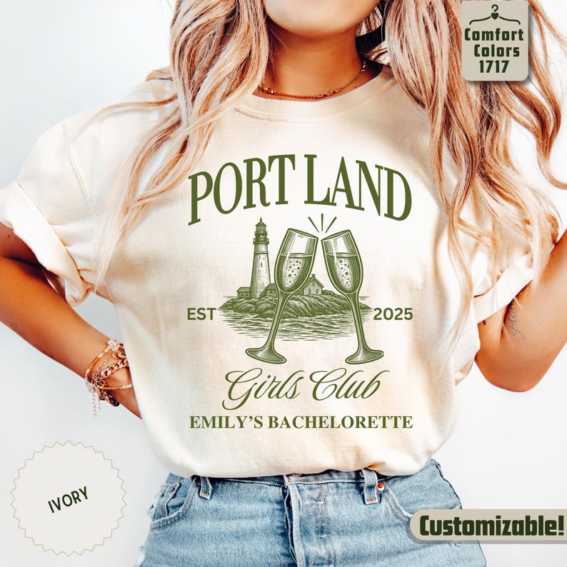 May include: An ivory t-shirt featuring a dark green graphic print with the text PORT LAND, EST 2025, Girls Club, and EMILY'S BACHELORETTE. The design includes an illustration of a lighthouse on a rocky shore and two clinking champagne flutes.