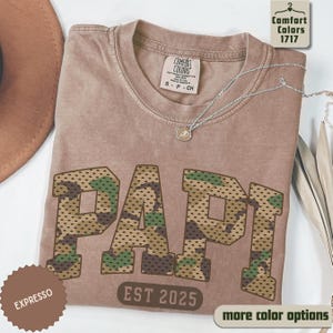 May include: A tan Comfort Colors t-shirt with the word "PAPI" in large, camouflage-patterned letters. The text "EST 2025" is below the word. A silver necklace with a pendant is on the shirt. The shirt is folded.