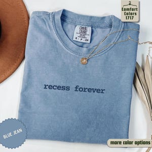 May include: A blue jean Comfort Colors t-shirt with the phrase "recess forever" printed on the front. A gold necklace with a pendant rests on the shirt. The shirt is a size small, petite, or child's size.