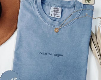 Może przedstawiać: Niebieska koszulka Comfort Colors z napisem "born to argue" prostą czcionką. Złoty naszyjnik z wisiorkiem spoczywa na koszulce. Koszulka ma okrągły dekolt i krótkie rękawy. Etykieta głosi "Comfort Colors 1717".
