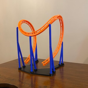 Könnte beinhalten: Ein Miniatur-Achterbahnmodell mit einer orangefarbenen Strecke und blauen Stützsäulen. Die Strecke weist eine Schleife und einen Drop auf, die auf einer schwarzen Basis angeordnet sind. Das Modell simuliert eine aufregende Vergnügungsparkfahrt.